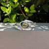 A polished silver-toned open cuff ring with a central oval green stone, resting on a light gray ledge with green foliage in the background.