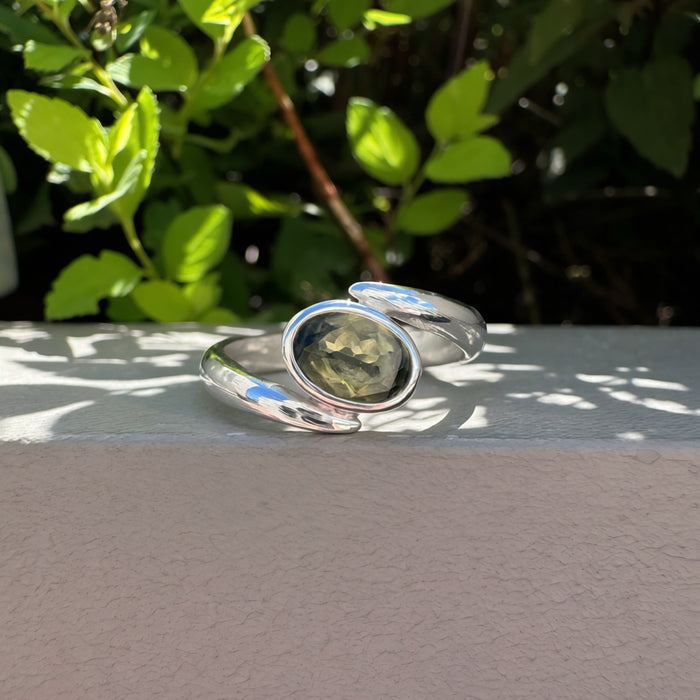 A polished silver-toned open cuff ring with a central oval green stone, resting on a light gray ledge with green foliage in the background.