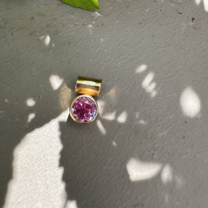 A gold-toned pendant with a round faceted pink gemstone, resting on a textured gray concrete surface with dappled sunlight and green leaves in the background.