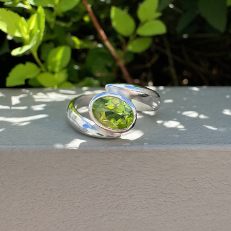 A silver wrap ring with a oval green gemstone, resting on a light gray textured surface with green foliage in the background.