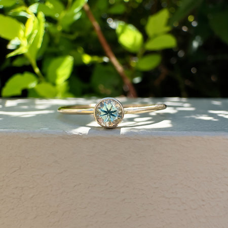 A gold ring with a round bezel-set light green gemstone, resting on a textured beige surface with a blurred green garden background.