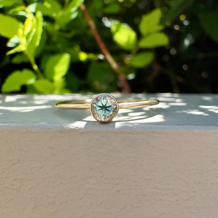 A gold ring with a round bezel-set light green gemstone, resting on a textured beige surface with a blurred green garden background.