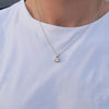 A close-up of a person wearing a delicate silver chain necklace with a small bezel-set clear round gemstone pendant, styled with a white crew-neck t-shirt.