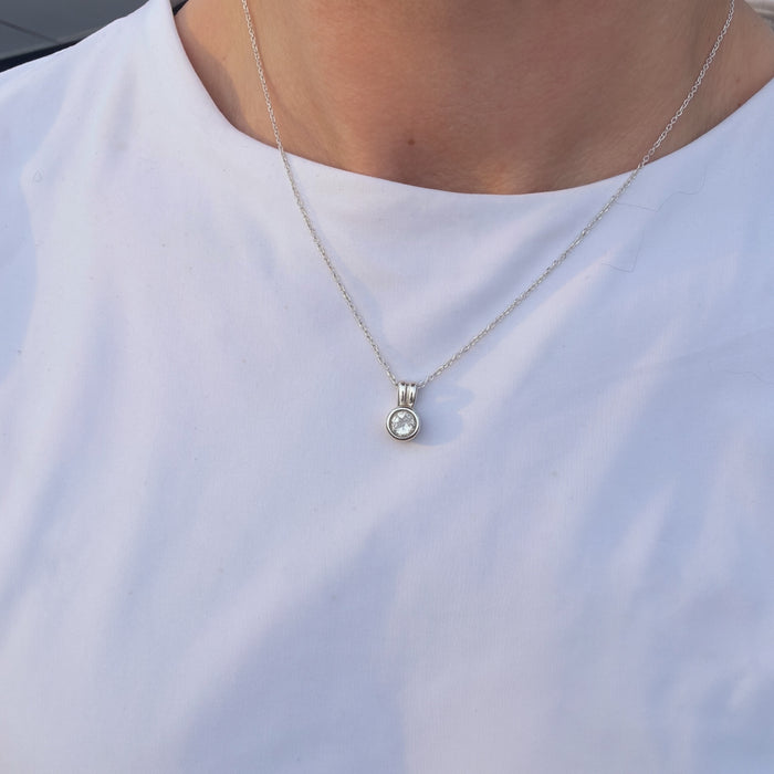 A close-up of a person wearing a delicate silver chain necklace with a small bezel-set clear round gemstone pendant, styled with a white crew-neck t-shirt.
