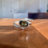 A sterling silver split-band ring with an oval bezel-set olive green gemstone, resting on a polished wooden tabletop with a blurred indoor background.