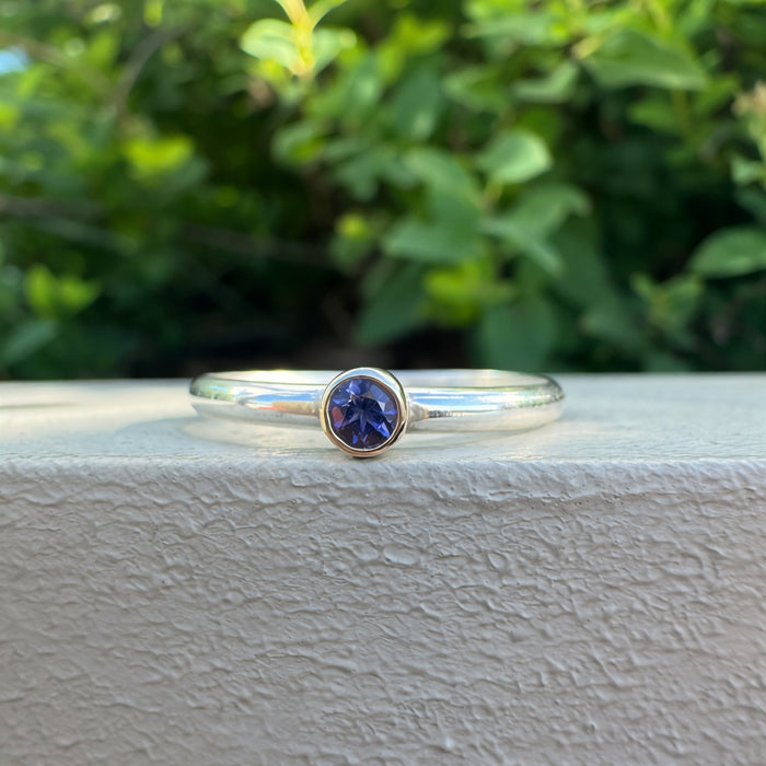 A thin silver band ring with a bezel-set round blue gemstone, resting on a textured white stone ledge with a blurred green garden background.