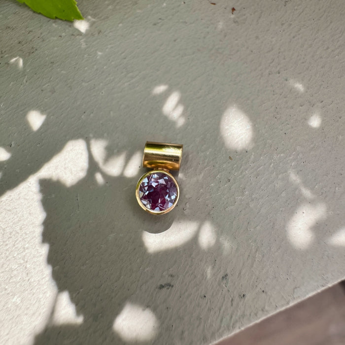 A gold bail pendant with a round purple gemstone, resting on a gray concrete surface with dappled sunlight and leaf shadows.