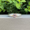 A silver band ring with a round pink gemstone in a bezel setting, resting on a textured gray surface with green foliage in the background.
