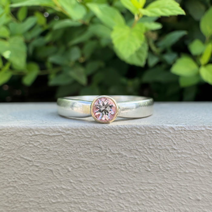 A silver band ring with a round pink gemstone in a bezel setting, resting on a textured gray surface with green foliage in the background.