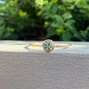 A delicate gold ring with a round-cut light green gemstone in a bezel setting, resting on the edge of a textured white wall with a soft-focus background of green foliage.