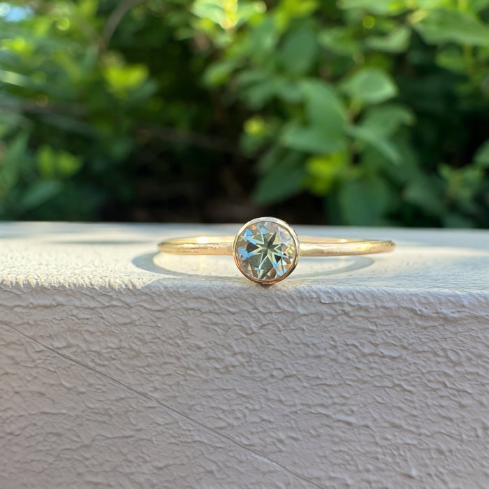 A delicate gold ring with a round-cut light green gemstone in a bezel setting, resting on the edge of a textured white wall with a soft-focus background of green foliage.