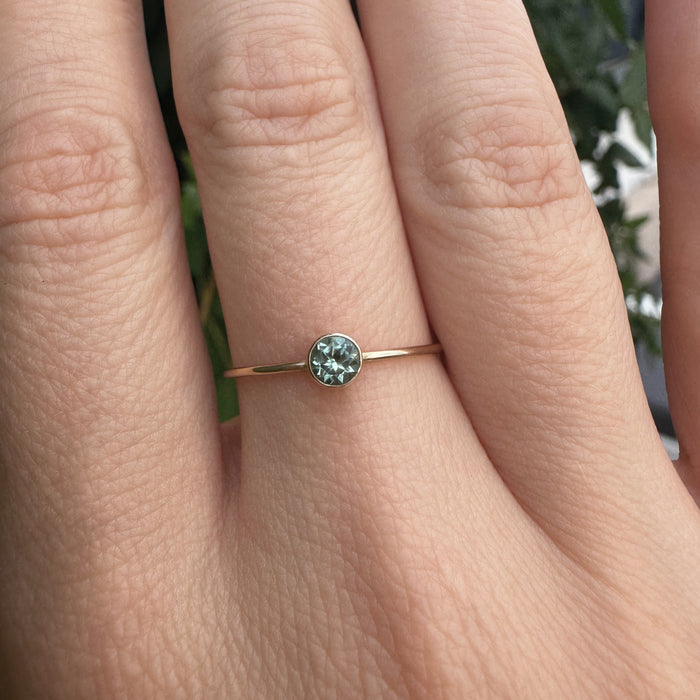 A delicate gold ring with a small round green gemstone set on a person's ring finger, shown in a close-up shot with a blurred background of green foliage.
