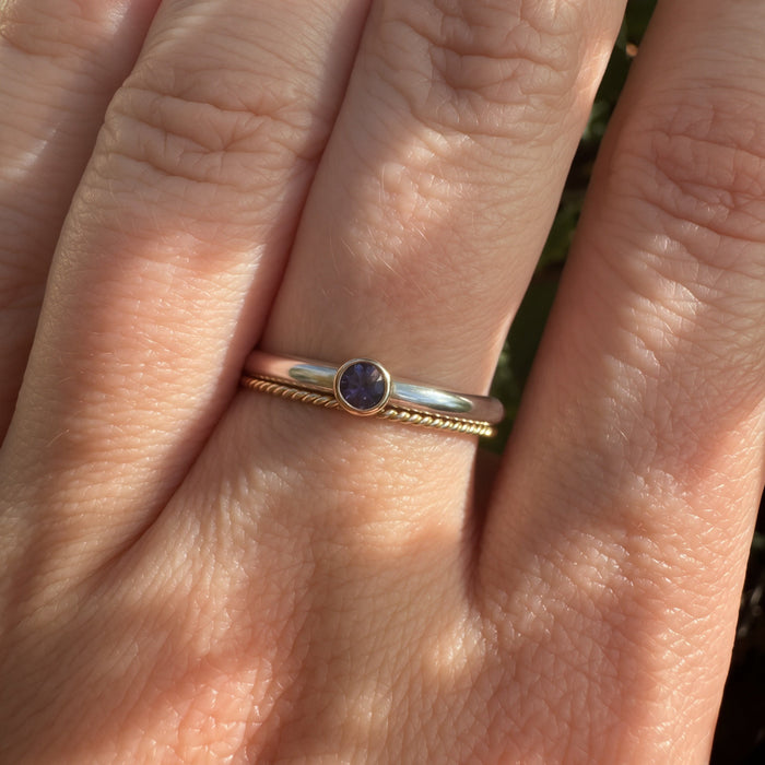 A close-up of a hand wearing a delicate ring with a small round blue gemstone and a twisted gold band.