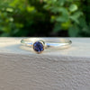 A silver ring with a bezel-set round blue gemstone, resting on a textured white surface with a blurred green garden background.