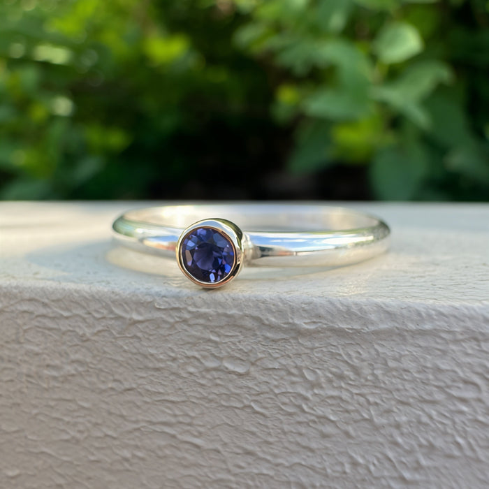 A silver ring with a bezel-set round blue gemstone, resting on a textured white surface with a blurred green garden background.
