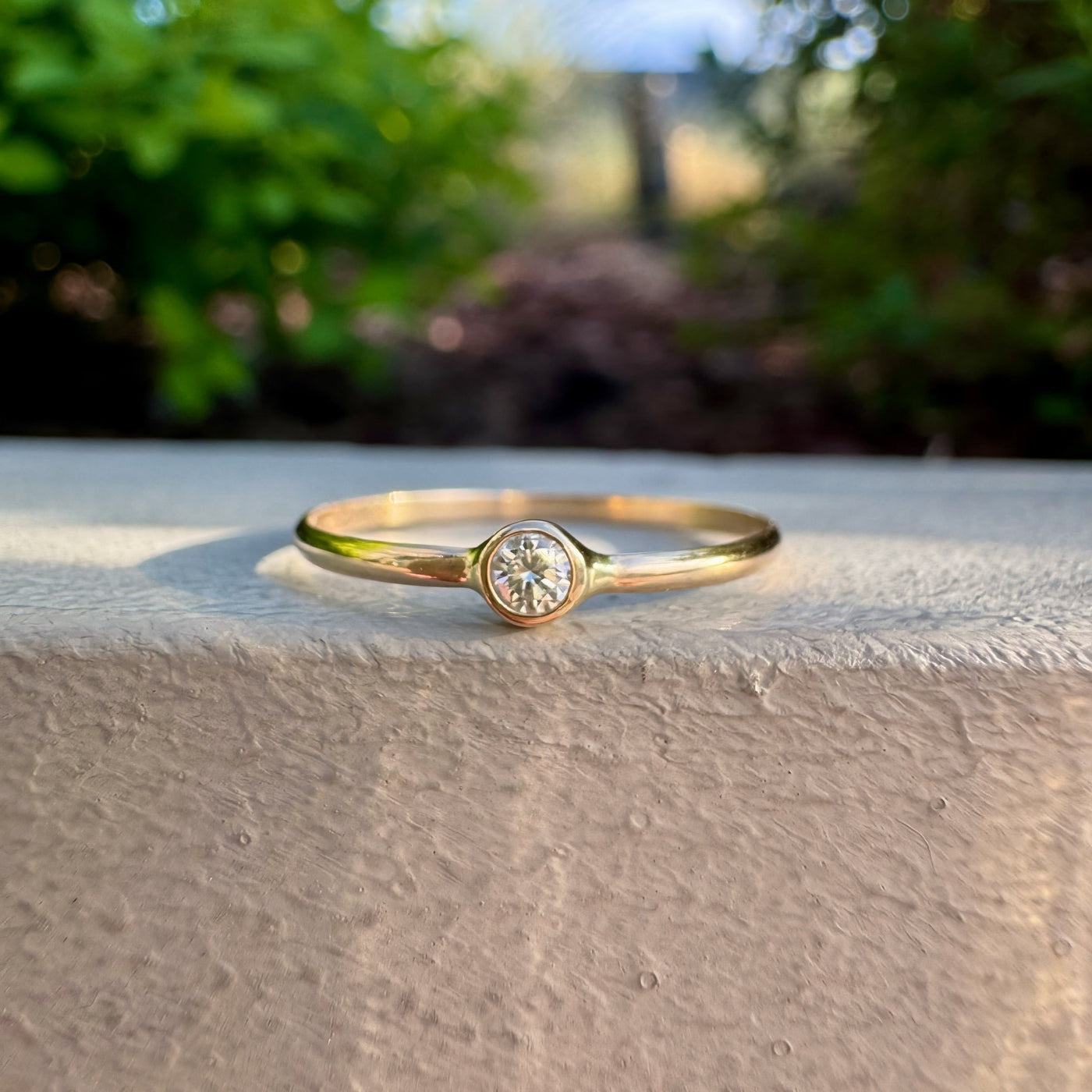 A delicate yellow gold ring with a single round-cut clear gemstone, resting on a textured beige stone ledge with a blurred green garden background.