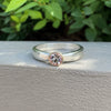 A silver band ring with a bezel-set round clear gemstone and a rose gold accent ring, resting on a textured light gray surface with a blurred green leafy background.