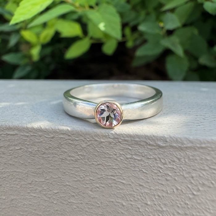 A silver band ring with a bezel-set round clear gemstone and a rose gold accent ring, resting on a textured light gray surface with a blurred green leafy background.