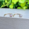 A pair of 14k gold open cuff earrings with round opal stones, displayed on a textured gray ledge with green foliage in the background.