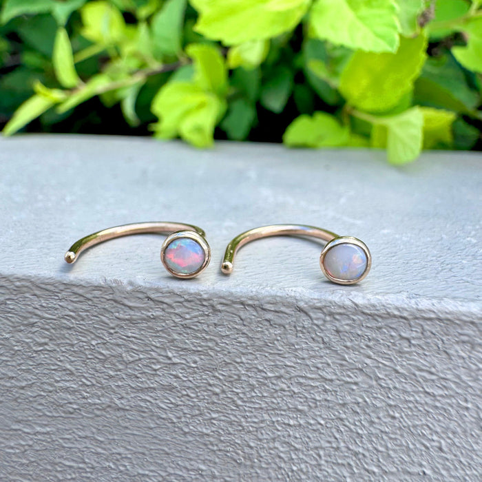 A pair of 14k gold open cuff earrings with round opal stones, displayed on a textured gray ledge with green foliage in the background.