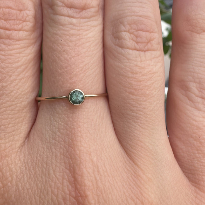 A close-up of a hand wearing a delicate gold ring with a small round green gemstone.