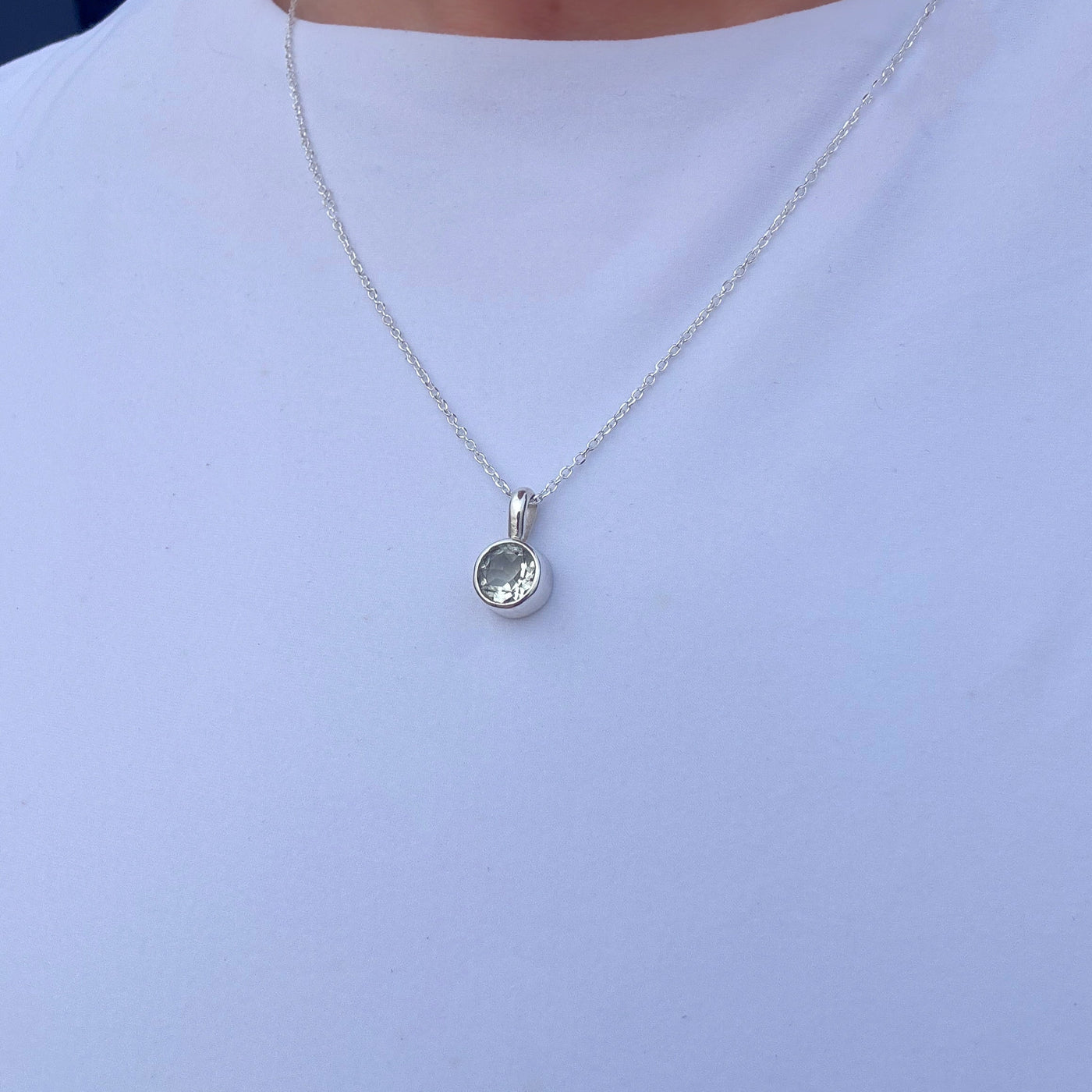A silver chain necklace with a small round bezel-set clear gemstone pendant, shown worn on a person's neck over a white t-shirt.