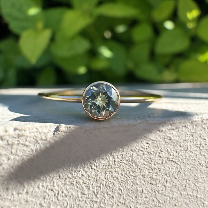 A delicate gold ring with a bezel-set round green gemstone, resting on a textured light gray stone surface with a blurred green foliage background.