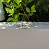A silver ring with an oval green gemstone, resting on a light gray concrete ledge with green foliage in the background.