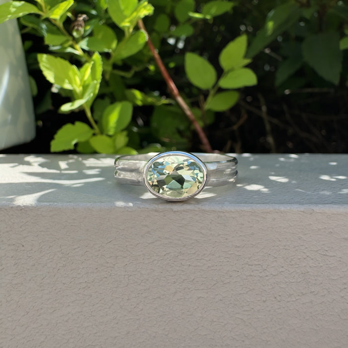 A silver ring with an oval green gemstone, resting on a light gray concrete ledge with green foliage in the background.