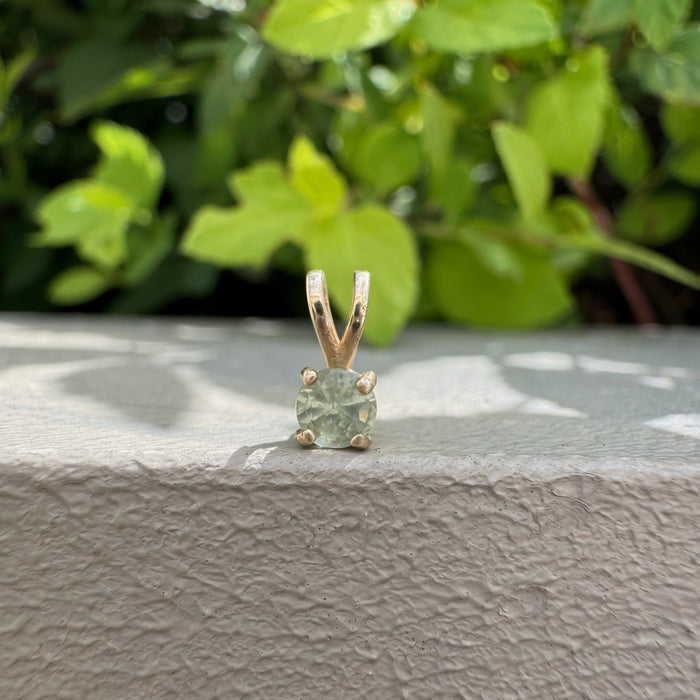 A gold four-prong pendant with a light green gemstone, resting on a textured gray stone ledge with a blurred background of green foliage.