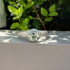 A silver ring with a round bezel-set green gemstone, resting on a light gray textured surface with green foliage in the background.