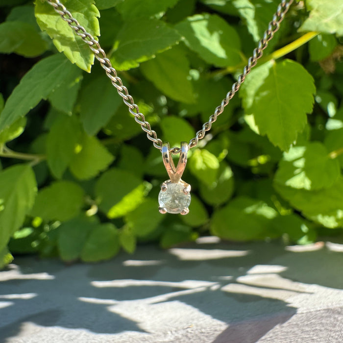 A delicate gold chain necklace with a small gold pendant holding a light green faceted gemstone, photographed outdoors against green leaves and sunlight.