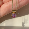 A gold necklace with a bezel-set round pink gemstone pendant, held in a person's hand against a light background.