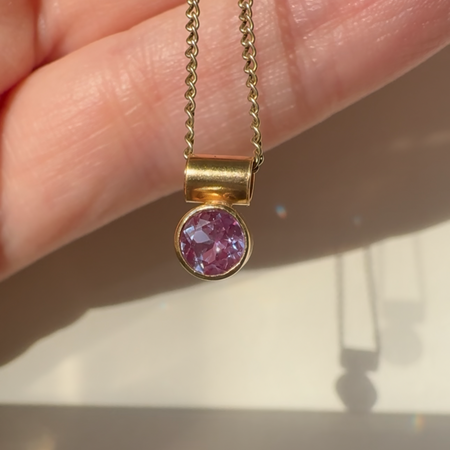 A gold necklace with a bezel-set round pink gemstone pendant, held in a person's hand against a light background.
