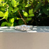 A silver ring with a large oval clear gemstone, resting on a light beige stone ledge with green foliage in the background.