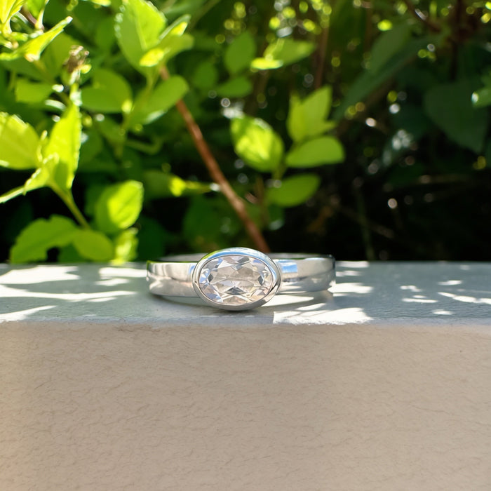 A silver ring with a large oval clear gemstone, resting on a light beige stone ledge with green foliage in the background.