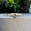 A gold solitaire engagement ring with a round-cut clear gemstone, resting on a textured white surface with a blurred green garden background.