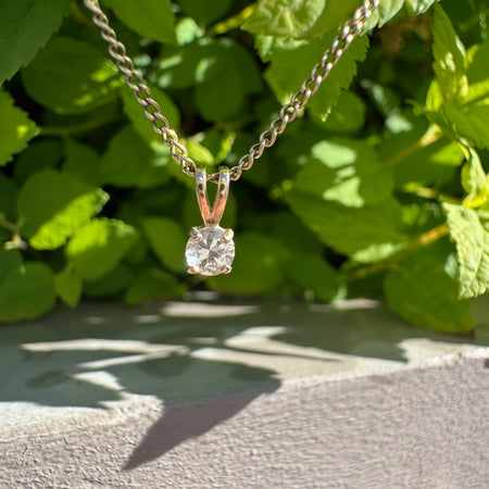 A gold chain necklace with a yellow gold pendant featuring a clear round-cut gemstone, photographed outdoors against a background of green leaves and a gray wall.