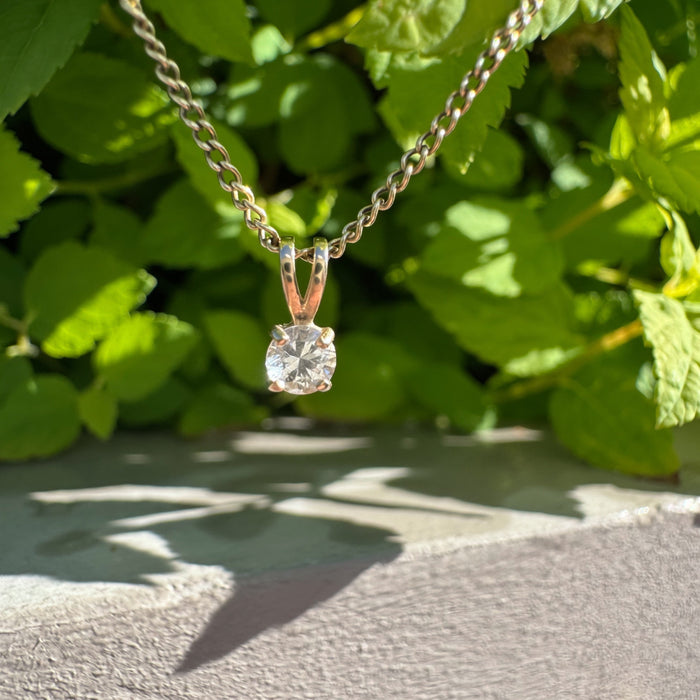 A gold chain necklace with a yellow gold pendant featuring a clear round-cut gemstone, photographed outdoors against a background of green leaves and a gray wall.