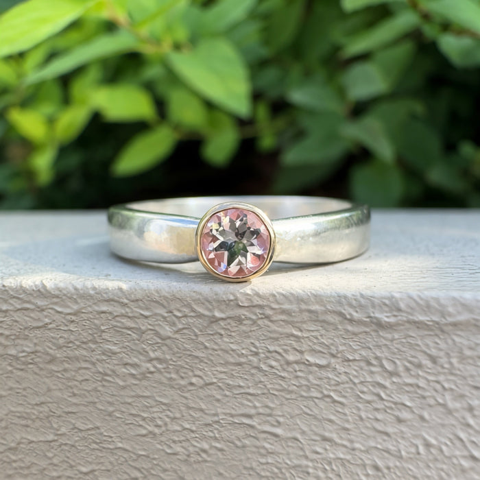 A silver ring with a round bezel-set pink gemstone, resting on a textured light gray surface with a blurred background of green leaves.
