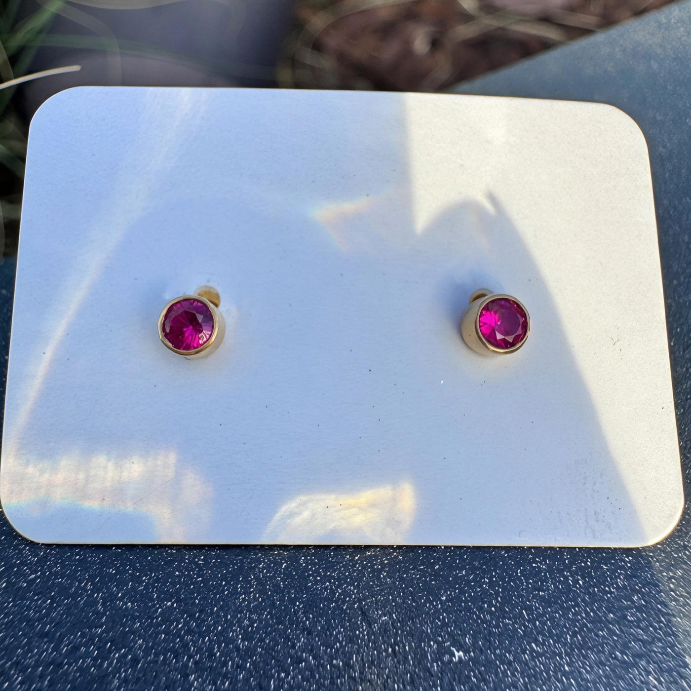 A pair of small gold stud earrings with round magenta-pink gemstones, displayed on a white card against a dark textured surface with a blurred outdoor background.