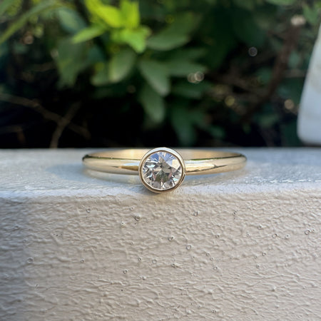 A yellow gold solitaire ring with a round-cut clear gemstone, resting on a textured white surface with a blurred green garden background.
