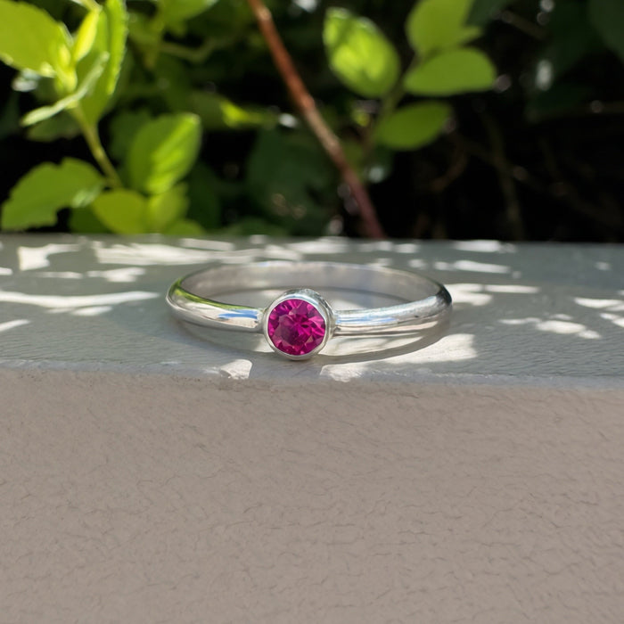 A thin silver band ring with a round pink gemstone, resting on a light beige textured surface with a background of green foliage.