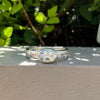 A silver band ring with a oval  clear gemstone, resting on a textured beige wall with green foliage in the background.