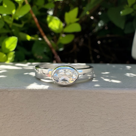 A silver band ring with a oval  clear gemstone, resting on a textured beige wall with green foliage in the background.