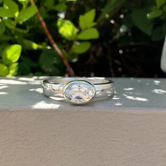 A silver band ring with a oval  clear gemstone, resting on a textured beige wall with green foliage in the background.