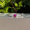 A silver ring with a round pink gemstone in a bezel setting, resting on a light beige textured surface with green foliage in the background.