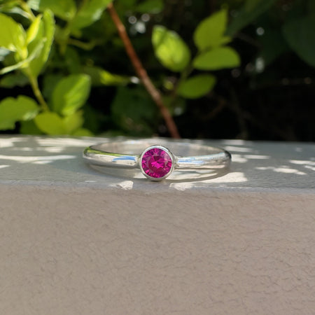 A silver ring with a round pink gemstone in a bezel setting, resting on a light beige textured surface with green foliage in the background.