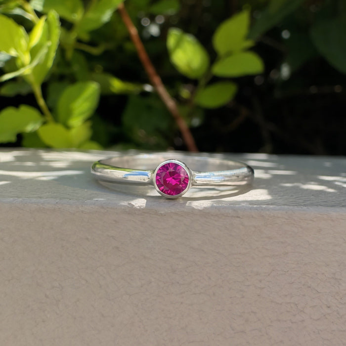 A silver ring with a round pink gemstone in a bezel setting, resting on a light beige textured surface with green foliage in the background.