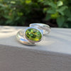 A silver ring with an oval green faceted gemstone, resting on a textured light gray surface with a blurred green garden background.